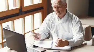Serious gray haired aged man in white shirt sitting at table and browsing laptop while checking notes in modern office